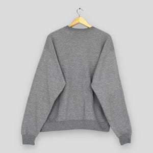 Puede incluir: Una sudadera gris jaspeada cuelga de una percha de madera. La sudadera de cuello redondo tiene mangas largas y pu&ntilde;os y dobladillo acanalados. La parte trasera de la sudadera es visible, mostrando un dise&ntilde;o sencillo e informal.