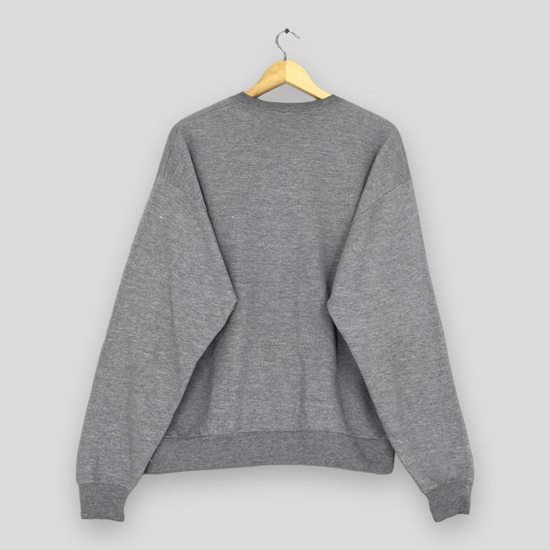Puede incluir: Una sudadera gris jaspeada cuelga de una percha de madera. La sudadera de cuello redondo tiene mangas largas y pu&ntilde;os y dobladillo acanalados. La parte trasera de la sudadera es visible, mostrando un dise&ntilde;o sencillo e informal.