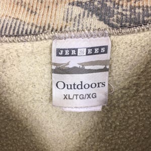 May include: Close-up of a tag from a Jerzees sweatshirt. The tag reads "Outdoors" in a bold font, with the size XL/TG/XG below. The background of the sweatshirt is a camouflage pattern in shades of brown, tan, and gray.