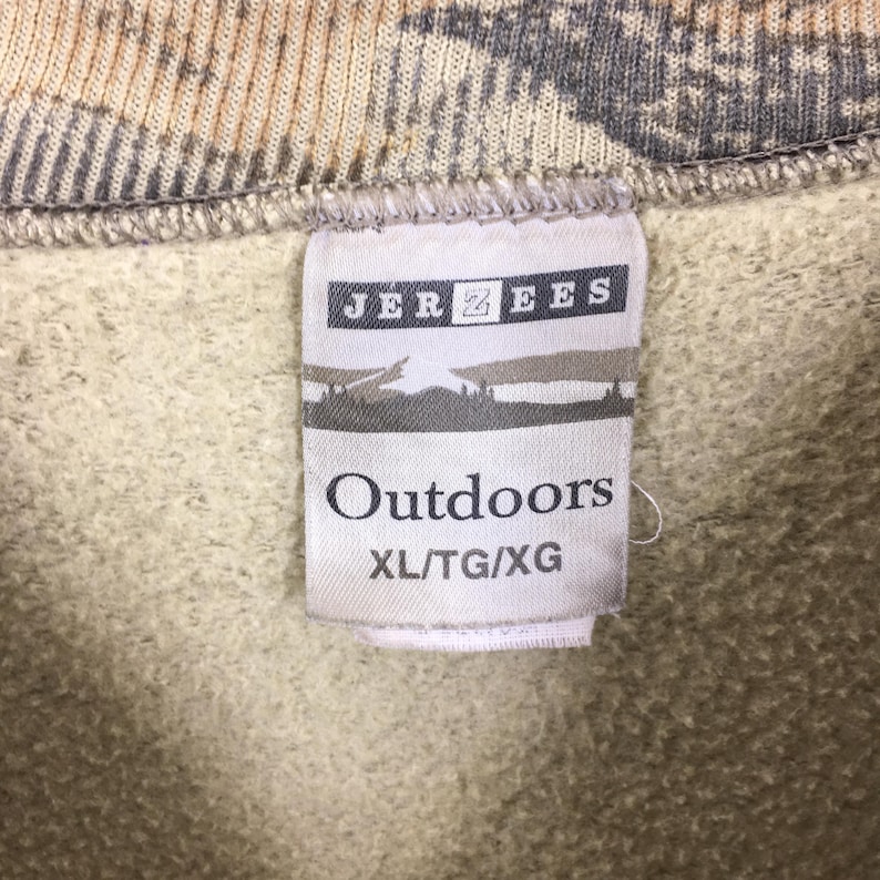 May include: Close-up of a tag from a Jerzees sweatshirt. The tag reads "Outdoors" in a bold font, with the size XL/TG/XG below. The background of the sweatshirt is a camouflage pattern in shades of brown, tan, and gray.