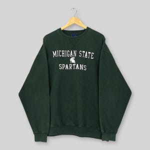 Puede incluir: Sudadera verde oscuro con las palabras "MICHIGAN STATE" y "SPARTANS" en blanco, junto con un gráfico de un casco espartano. La sudadera está colgada de una percha de madera. Es un estilo clásico de cuello redondo.