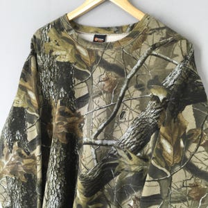 May include: A camouflage-patterned sweatshirt in shades of brown, green, and tan, resembling a forest scene. The crew neck sweatshirt is hanging on a wooden hanger. The fabric features a detailed print of branches and leaves.
