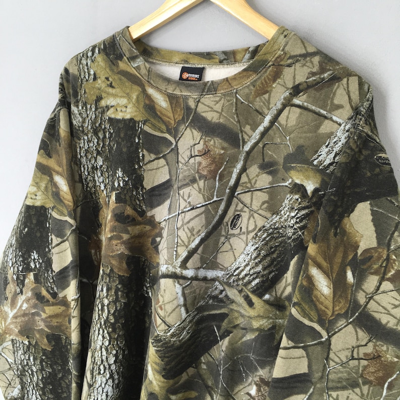 May include: A camouflage-patterned sweatshirt in shades of brown, green, and tan, resembling a forest scene. The crew neck sweatshirt is hanging on a wooden hanger. The fabric features a detailed print of branches and leaves.