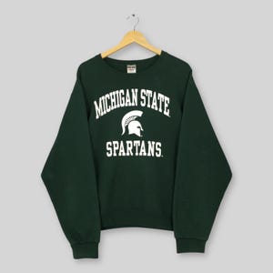 Puede incluir: Una sudadera verde oscuro con las palabras "MICHIGAN STATE" en blanco en la parte superior y "SPARTANS" debajo. Un casco espartano blanco está entre las palabras. La sudadera está colgada de una percha de madera. Es un estilo clásico de cuello redondo.