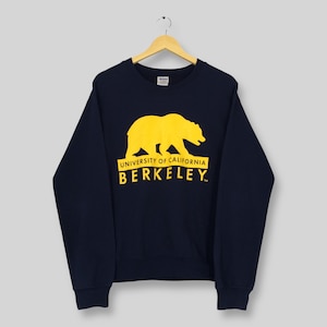 Peut inclure: Sweat-shirt bleu marine avec un ours jaune et l'inscription "UNIVERSITY OF CALIFORNIA BERKELEY". Le sweat-shirt est suspendu à un cintre en bois.