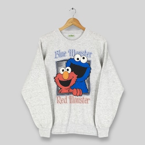 Puede incluir: Sudadera gris claro con el texto "Blue Monster" y "Red Monster" y un gráfico de Elmo y Cookie Monster de Barrio Sésamo. La sudadera tiene mangas largas y cuello redondo.