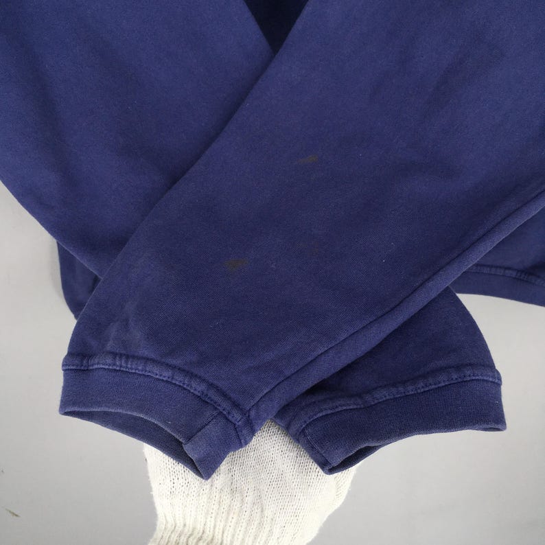 May include: Close-up of a navy blue sweatshirt sleeve. The sleeve is cuffed at the wrist and shows some discoloration. The fabric appears to be a soft, knit material. The image is well-lit, highlighting the texture and colour of the garment.