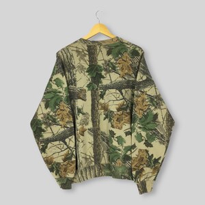 May include: A long-sleeved sweatshirt with a camouflage pattern of tree branches and leaves in shades of brown, green, and tan. The sweatshirt is hanging on a wooden hanger against a light gray background. The overall design gives a natural, outdoorsy feel.