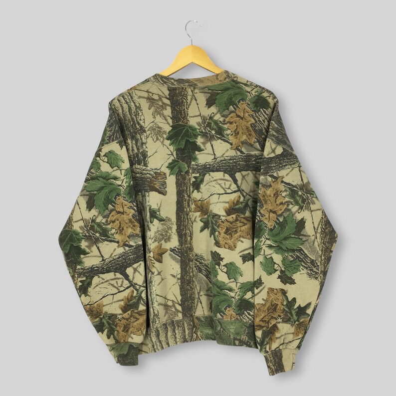 May include: A long-sleeved sweatshirt with a camouflage pattern of tree branches and leaves in shades of brown, green, and tan. The sweatshirt is hanging on a wooden hanger against a light gray background. The overall design gives a natural, outdoorsy feel.