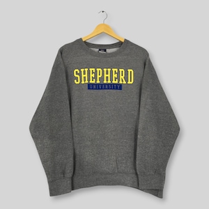 Peut inclure: Un sweat-shirt gris avec l'inscription "SHEPHERD UNIVERSITY" en lettres jaunes au-dessus d'un rectangle bleu. Le sweat-shirt est suspendu à un cintre en bois sur un fond gris clair.