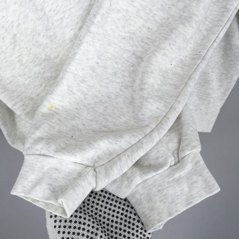 Peut inclure: Gros plan sur une manche de sweat-shirt gris clair avec un poignet c&ocirc;tel&eacute;. Le tissu a une texture mouchet&eacute;e et une petite tache jaune. La manche est repli&eacute;e, r&eacute;v&eacute;lant une partie d'un v&ecirc;tement &agrave; motifs noir et blanc en dessous.