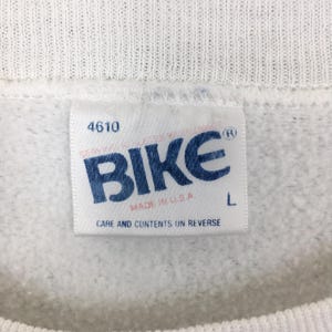May include: Close-up of a white sweatshirt tag with the word "BIKE" in blue block letters. The tag also includes the text "MADE IN U.S.A." and "CARE AND CONTENTS ON REVERSE". The size is L.
