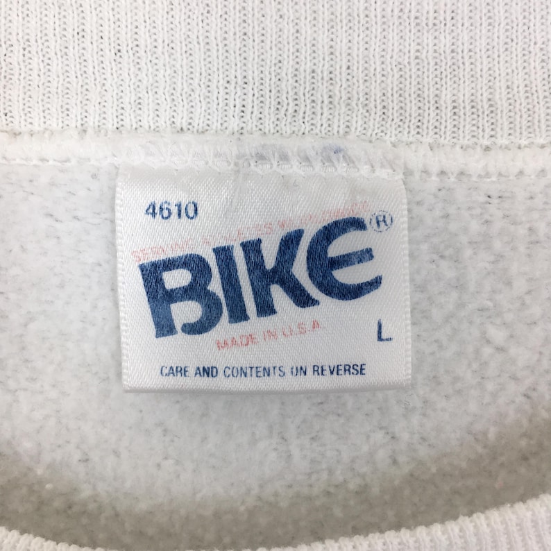 May include: Close-up of a white sweatshirt tag with the word "BIKE" in blue block letters. The tag also includes the text "MADE IN U.S.A." and "CARE AND CONTENTS ON REVERSE". The size is L.