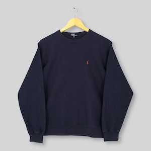 May include: A navy blue sweatshirt with a crew neck and long sleeves. The sweatshirt features a small red logo on the chest. The garment is hanging on a wooden hanger against a light gray background.