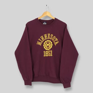Könnte beinhalten: Burgunderfarbenes Sweatshirt mit dem Wort "MINNESOTA" in gelben Buchstaben auf der Brust. Darunter ein gelbes Emblem und die Zahl "1851". Das Sweatshirt hängt an einem Holzkleiderbügel.