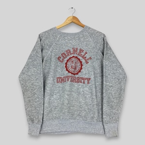 May include: A heather gray long-sleeve sweatshirt featuring a crew neckline and raglan sleeves. The front displays the text CORNELL UNIVERSITY in red lettering arched above and below a circular red emblem containing a crest and additional text.
