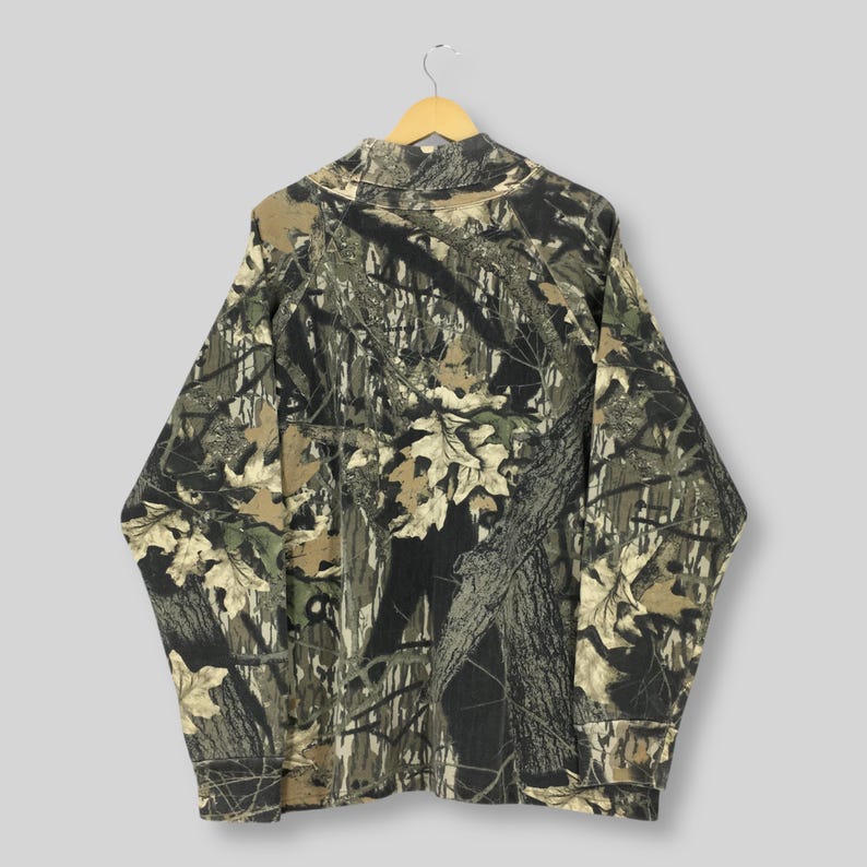 May include: A long-sleeved camouflage shirt with a mock neck, hanging from a wooden hanger. The shirt features a pattern of brown, green, and tan leaves and tree branches. The shirt is made of a soft, comfortable material.