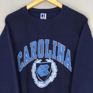 May include: Navy blue sweatshirt with "CAROLINA" arched across the chest in light blue. Below the text is a light blue shield with a "NC" logo, surrounded by a white wreath. The sweatshirt has a crew neck and long sleeves.