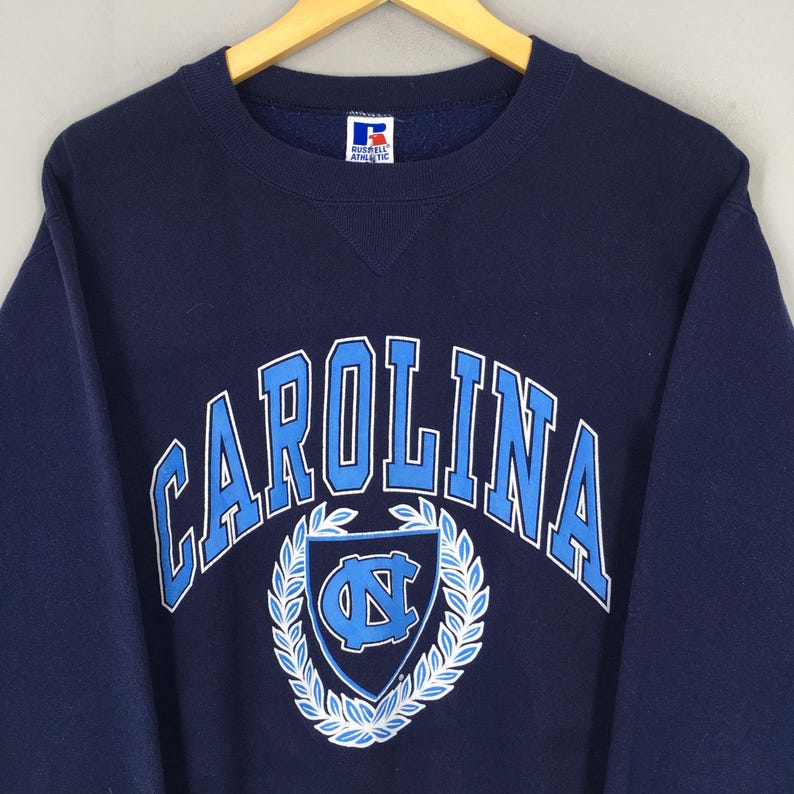 May include: Navy blue sweatshirt with "CAROLINA" arched across the chest in light blue. Below the text is a light blue shield with a "NC" logo, surrounded by a white wreath. The sweatshirt has a crew neck and long sleeves.