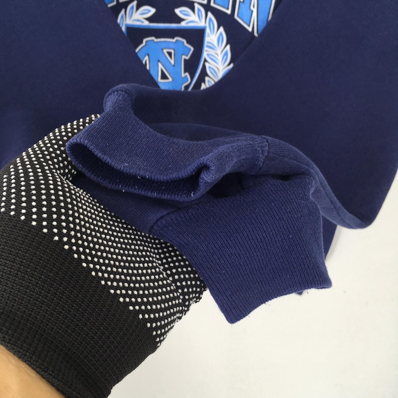 May include: Navy blue sweatshirt with a light blue emblem featuring a stylized "N" and the text "UNC". The sweatshirt has a ribbed cuff and a black and white patterned sleeve.