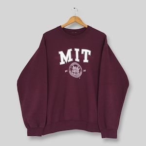 Könnte beinhalten: Ein kastanienbraunes Langarm-Sweatshirt mit Rundhalsausschnitt, das den weißen Schriftzug MIT in gebogener Schrift über einem kreisförmigen Siegel mit den Worten Massachusetts Institute of Technology und EST. 1861 auf der Brust zeigt.