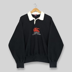 May include: A black long-sleeve rugby shirt featuring a white collar and a button placket. The chest displays an embroidered red logo and the text CANTERBURY OF NEW ZEALAND in white. The garment has ribbed cuffs and a ribbed hem, hanging on a hanger.