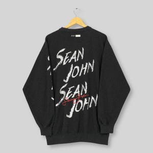 以下が含まれることがあります： 背面に白のスタイリッシュな文字で「SEAN JOHN」と書かれた濃いグレーのスウェットシャツ。ブランド名は3回繰り返され、中央のインスタンスは筆記体と赤インクで書かれています。スウェットシャツは木製のハンガーに吊るされています。