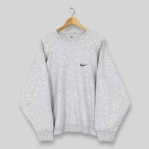 May include: A light grey Nike sweatshirt with a small black Nike swoosh logo on the chest. The long-sleeved crew neck sweatshirt is hanging on a wooden hanger. There are a few small yellow stains on the left sleeve.
