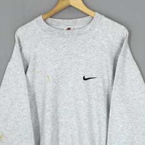 May include: A light grey crew neck sweatshirt with a small black Nike swoosh logo on the chest. The sweatshirt has a small Nike tag at the neckline and is hanging on a wooden hanger.