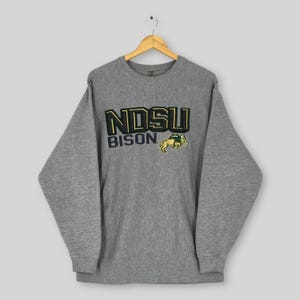 Może przedstawiać: Szara melanżowa bluza z napisem "NDSU BISON" w zielonych i czarnych literach. Żółto-zielone logo bizona znajduje się po prawej stronie napisu. Bluza wisi na drewnianym wieszaku.