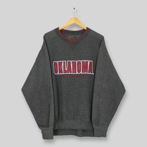 May include: A dark gray sweatshirt with a V-neck and red trim. The word "OKLAHOMA" is printed across the chest in white with a red outline. Below the word "OKLAHOMA" is the word "SOONERS" in a smaller font.