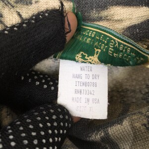 May include: Close-up of a camouflage-patterned garment with a green and white tag. The tag reads "WATER, HANG TO DRY, ITEM#0788, RN#73342, MADE IN USA, SIZE XL". The garment is held by a person wearing a black glove.