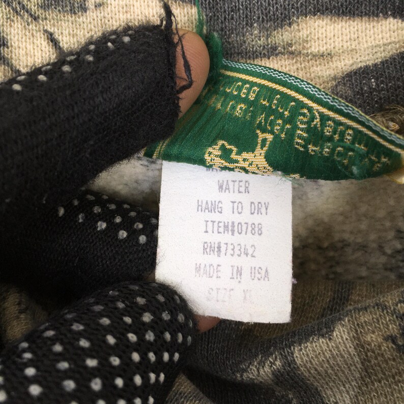 May include: Close-up of a camouflage-patterned garment with a green and white tag. The tag reads "WATER, HANG TO DRY, ITEM#0788, RN#73342, MADE IN USA, SIZE XL". The garment is held by a person wearing a black glove.