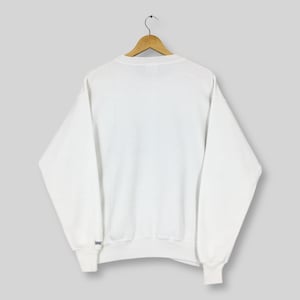 May include: A white crew neck sweatshirt with long sleeves, hanging on a wooden hanger. The cuff of the left sleeve has a small blue and white logo. The sweatshirt is made of a soft, comfortable material.