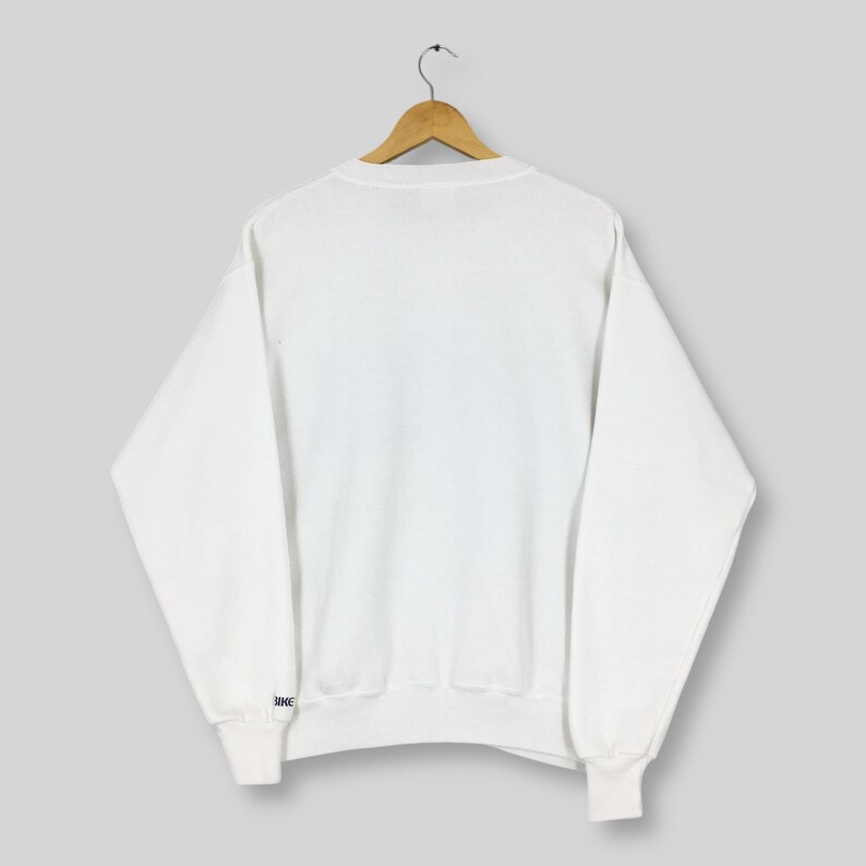 May include: A white crew neck sweatshirt with long sleeves, hanging on a wooden hanger. The cuff of the left sleeve has a small blue and white logo. The sweatshirt is made of a soft, comfortable material.