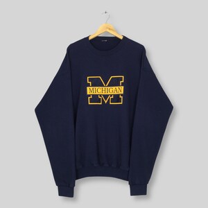 May include: Navy blue sweatshirt with a yellow "MICHIGAN" logo. The logo is a stylized "M" with the word "MICHIGAN" in a rectangular box. The sweatshirt is hanging on a wooden hanger.