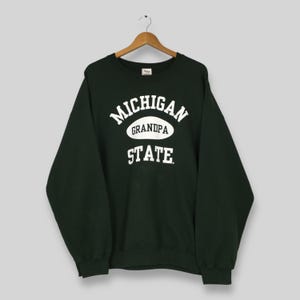 Puede incluir: Sudadera verde oscuro con las palabras "MICHIGAN GRANDPA STATE" en letras blancas. La sudadera está colgada de una percha de madera.