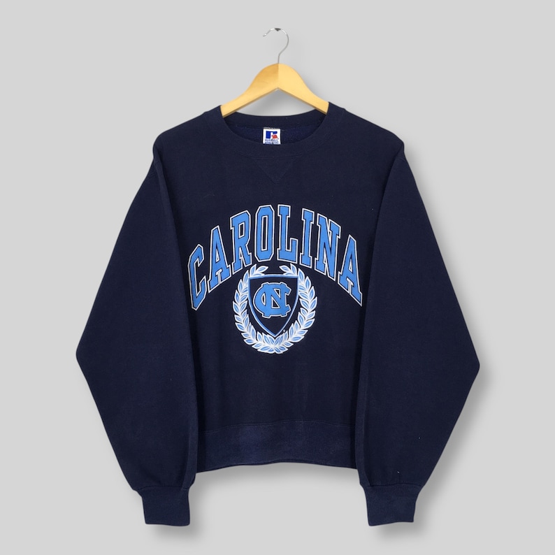 May include: Navy blue crewneck sweatshirt with "CAROLINA" arched across the chest in light blue lettering. Below the text is a light blue emblem with a wreath design. The sweatshirt is hanging on a wooden hanger.