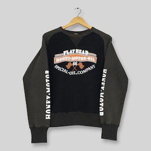 May include: A black and dark gray long-sleeve sweatshirt featuring a graphic on the chest that reads FLAT HEAD HONEY-MOTOR-OIL 66 SPECIAL-OIL, COMPANY with two checkered flags. The text HONEY-MOTOR is printed vertically on both sleeves.