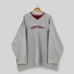May include: A gray sweatshirt with a V-neck collar and long sleeves. The word "GRUMPY" is embroidered in red across the chest. The collar has a red trim with the number "1937" visible.