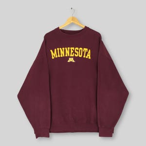Puede incluir: Una sudadera de color granate con cuello redondo y manga larga que presenta la palabra MINNESOTA en letras amarillas sobre el pecho, situada encima de un logotipo M amarillo. La prenda cuelga de una percha de madera sobre un fondo gris claro.