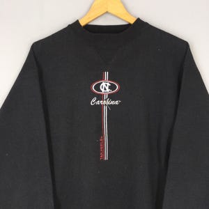 May include: Black sweatshirt with a crew neck and long sleeves. Features a logo with the text "Carolina" and "Tar Heels" in white and red. The logo is centered on the chest.