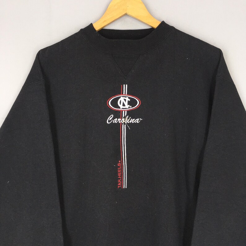 May include: Black sweatshirt with a crew neck and long sleeves. Features a logo with the text "Carolina" and "Tar Heels" in white and red. The logo is centered on the chest.