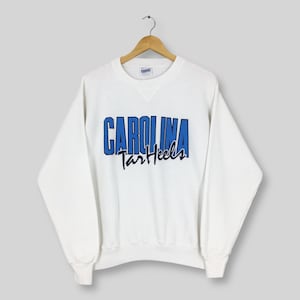 May include: White crew neck sweatshirt with the text "CAROLINA Tar Heels" in blue and navy lettering. The sweatshirt is hanging on a wooden hanger against a light gray background. The sweatshirt has long sleeves and a ribbed neckline.
