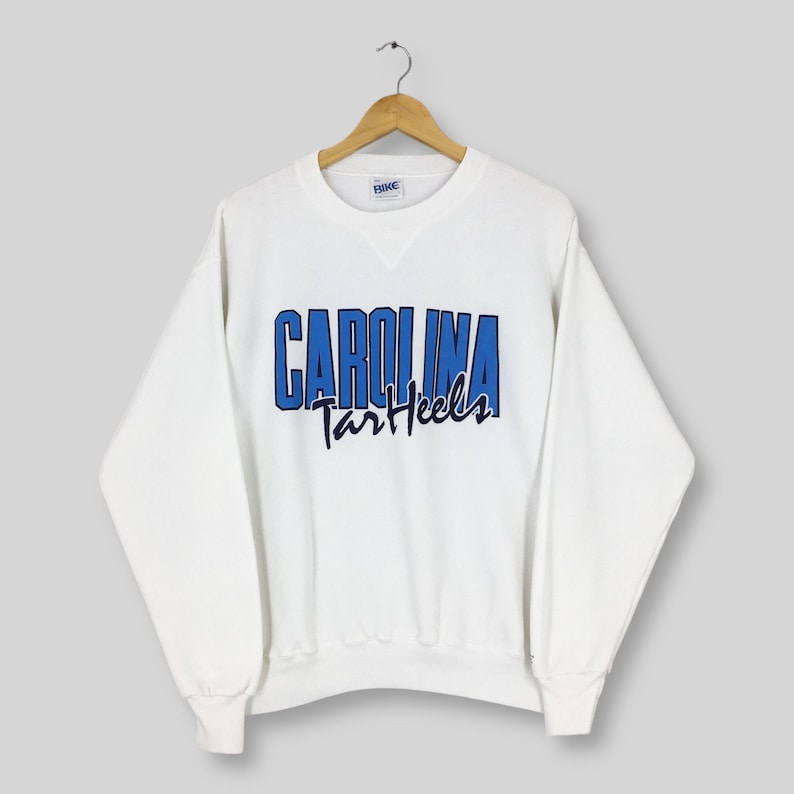 May include: White crew neck sweatshirt with the text "CAROLINA Tar Heels" in blue and navy lettering. The sweatshirt is hanging on a wooden hanger against a light gray background. The sweatshirt has long sleeves and a ribbed neckline.