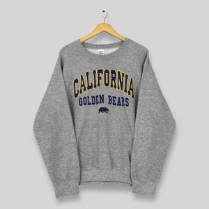 Könnte beinhalten: Ein graues Sweatshirt mit dem Wort "CALIFORNIA" in marineblauen und goldenen Buchstaben in Bogenform. Darunter steht der Text "GOLDEN BEARS" und eine marineblaue Bär-Grafik. Das Sweatshirt hängt an einem Holzkleiderbügel.