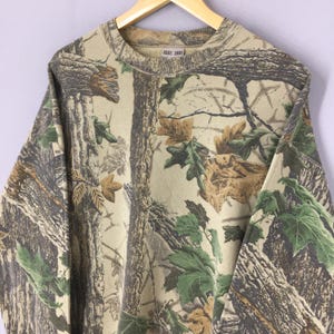 May include: A long-sleeved sweatshirt with a camouflage pattern. The design features a mix of brown, green, and tan colors, mimicking a forest environment. The sweatshirt has a crew neck and a relaxed fit, suitable for casual wear.