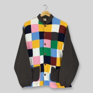 May include: A colourful button-up jacket with a patchwork design of squares in pink, yellow, blue, and navy. The sleeves are dark grey, and the jacket has two front pockets and wooden buttons. The collar is dark grey.