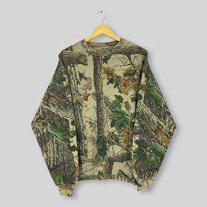May include: A camouflage-patterned sweatshirt hangs on a wooden hanger. The long-sleeved top features a realistic print of trees, branches, and leaves in shades of brown, green, and tan. The crew neck sweatshirt has ribbed cuffs and hem.