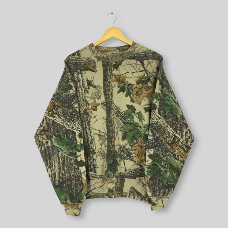 May include: A camouflage-patterned sweatshirt hangs on a wooden hanger. The long-sleeved top features a realistic print of trees, branches, and leaves in shades of brown, green, and tan. The crew neck sweatshirt has ribbed cuffs and hem.